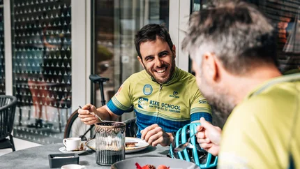 Two men in cycling gear eating and smiling at an outdoor table