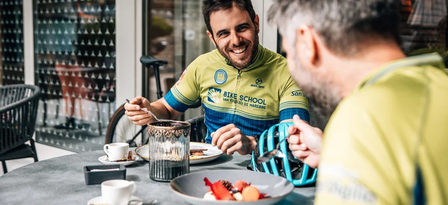 Zwei Männer in Fahrradkleidung essen und lachen an einem Außentisch