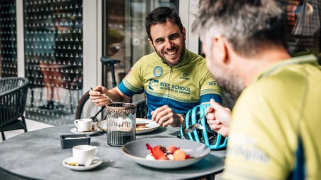 Zwei Männer in Fahrradkleidung essen und lachen an einem Außentisch