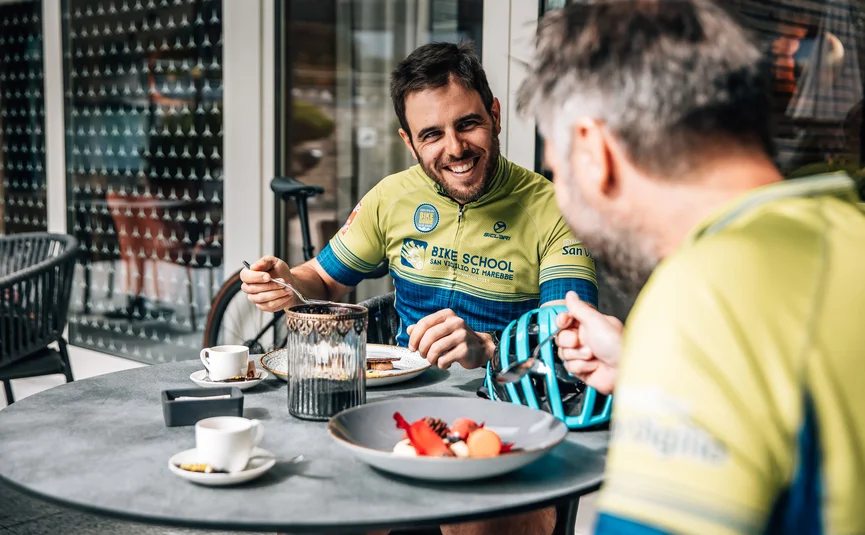 Zwei Männer in Fahrradkleidung essen und lachen an einem Außentisch