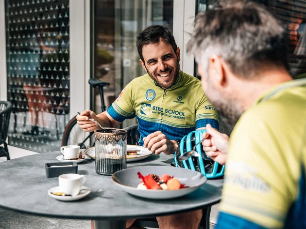 Zwei Männer in Fahrradkleidung essen und lachen an einem Außentisch