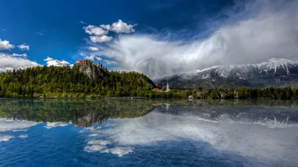 Burg auf bewaldetem Hügel mit See und Bergen bei bewölktem Himmel