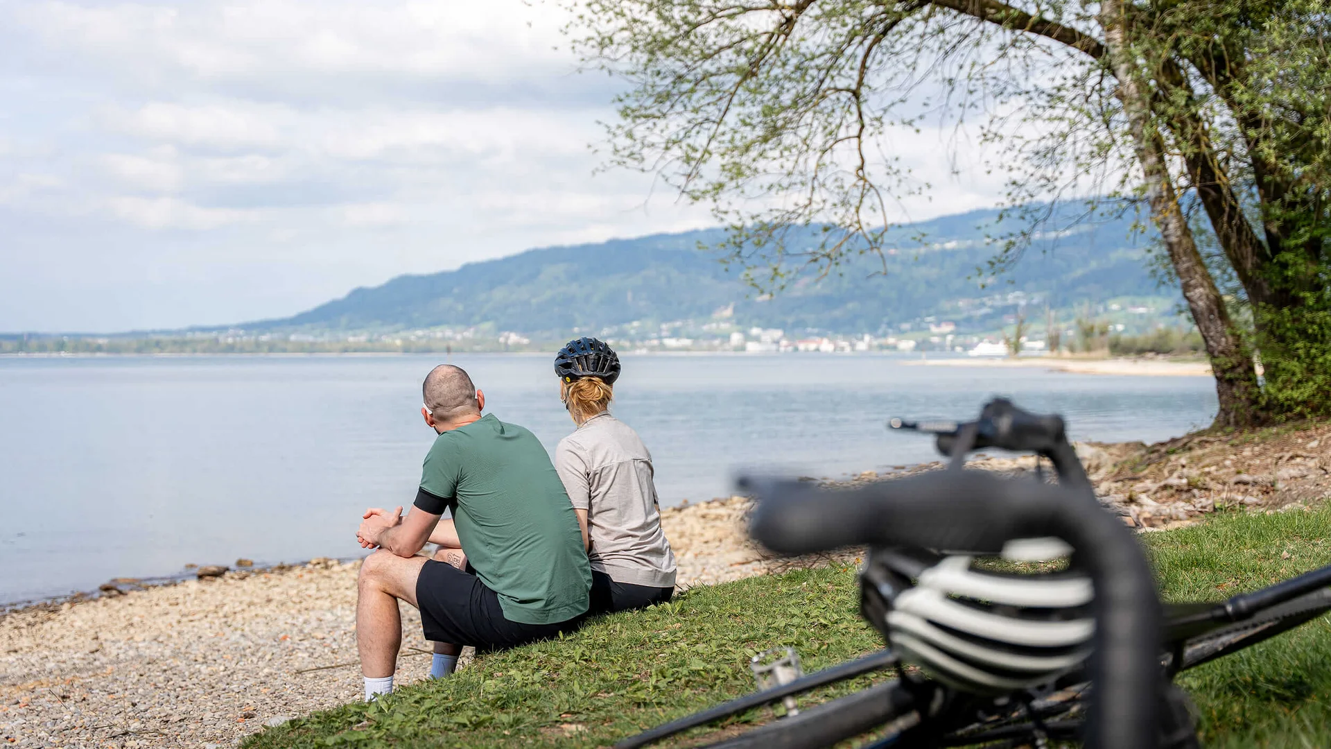 Grenzenlos graveln in Vorarlberg © Chris Gollhofer - Vorarlberg Tourismus Zwei Radfahrer sitzen am Seeufer und genießen die Aussicht auf das Wasser und die Berge