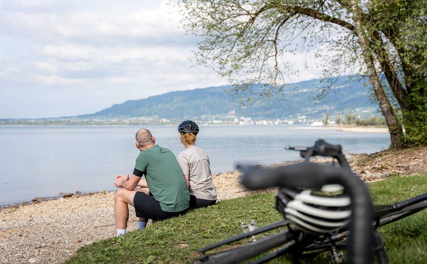 Zwei Radfahrer sitzen am Seeufer und genießen die Aussicht auf das Wasser und die Berge