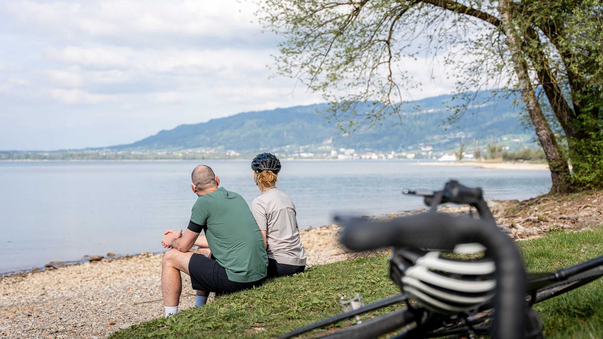 Zwei Radfahrer sitzen am Seeufer und genießen die Aussicht auf das Wasser und die Berge