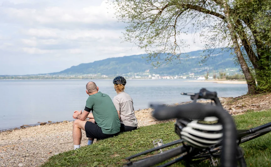 Two cyclists sitting by the lakeside enjoying the view of the water and mountains