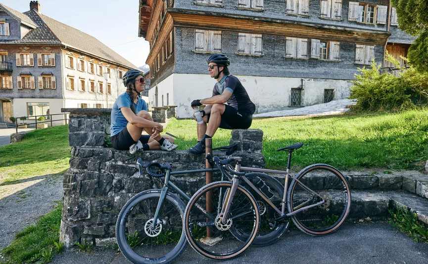 Two cyclists wearing helmets sitting on a stone wall near old buildings