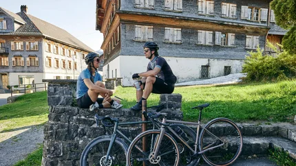 Zwei Radfahrer mit Helm sitzen auf einer Mauer vor historischen Gebäuden