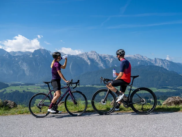 Zwei Radfahrer trinken Wasser mit Bergpanorama im Hintergrund