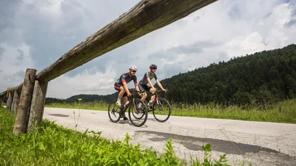 Two cyclists on country road next to green grass and forest under cloudy sky