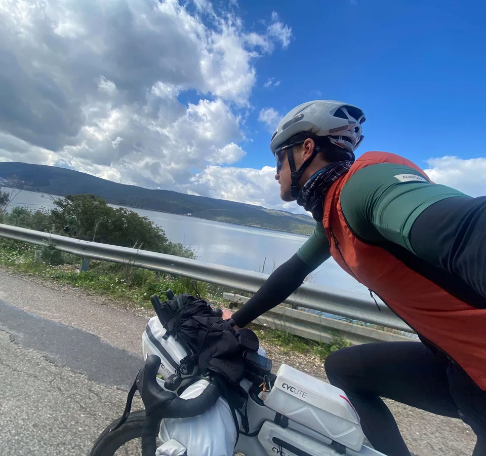 Bikepacking von Wien nach Athen © Jan Koller Radfahrer mit Helm und Ausrüstung fährt an einem See vorbei unter bewölktem Himmel
