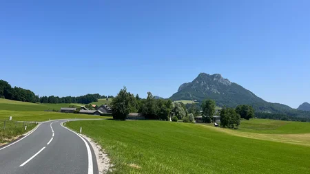 Drei Seen, eine Gravelrunde: Die 3-Seen-Tour ab Fuschl Landstraße führt durch grüne Wiesen zu einem bewaldeten Berg unter klarem blauen Himmel