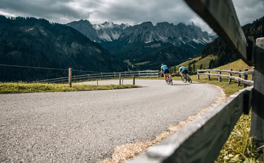Zwei Radfahrer fahren auf kurviger Bergstraße mit Zaun und bewölktem Himmel