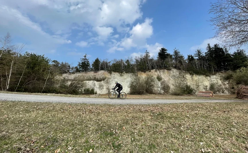 Radfahrer fährt auf Schotterweg vor bewaldetem Hang unter blauem Himmel