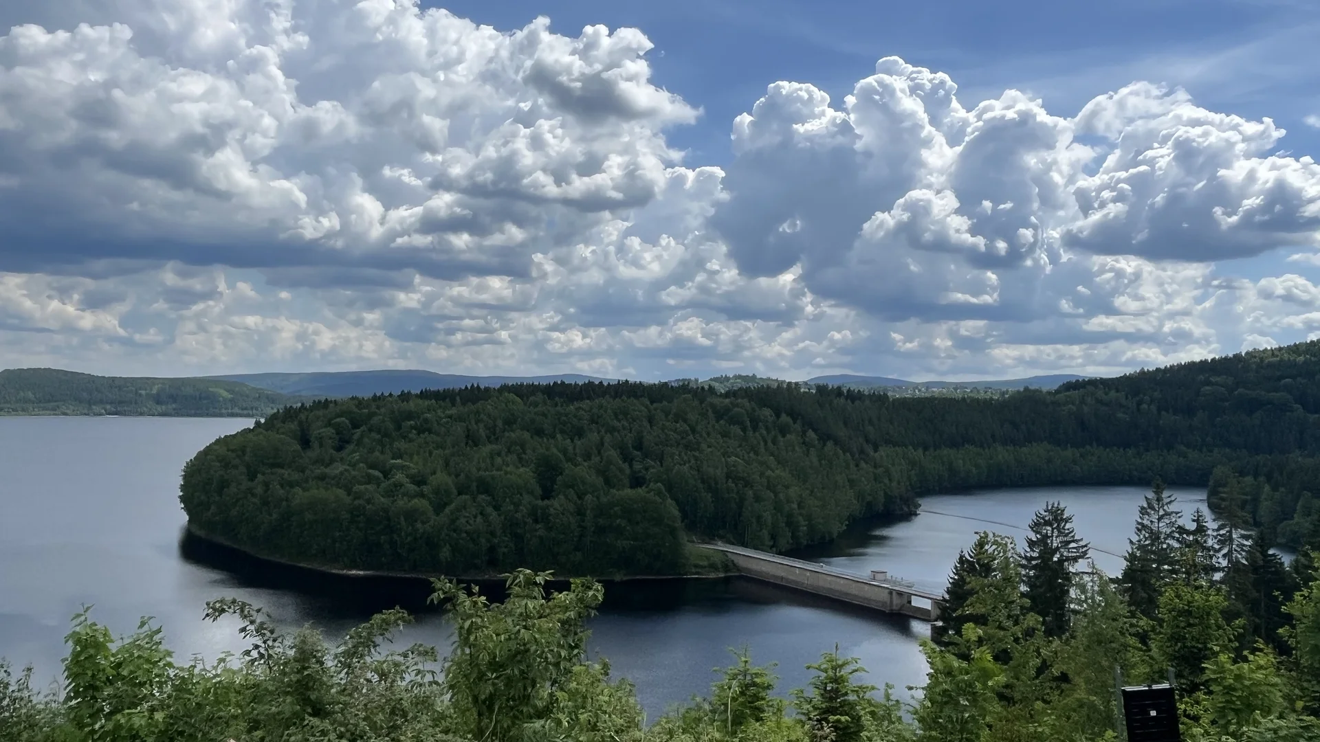 Landschaft mit See, Damm und bewaldetem Hügel unter bewölktem Himmel