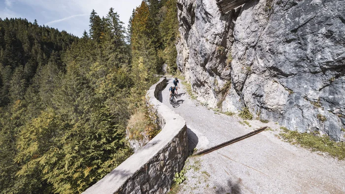 Zwei Radfahrer auf schmalem Bergweg neben Felswand und Wald