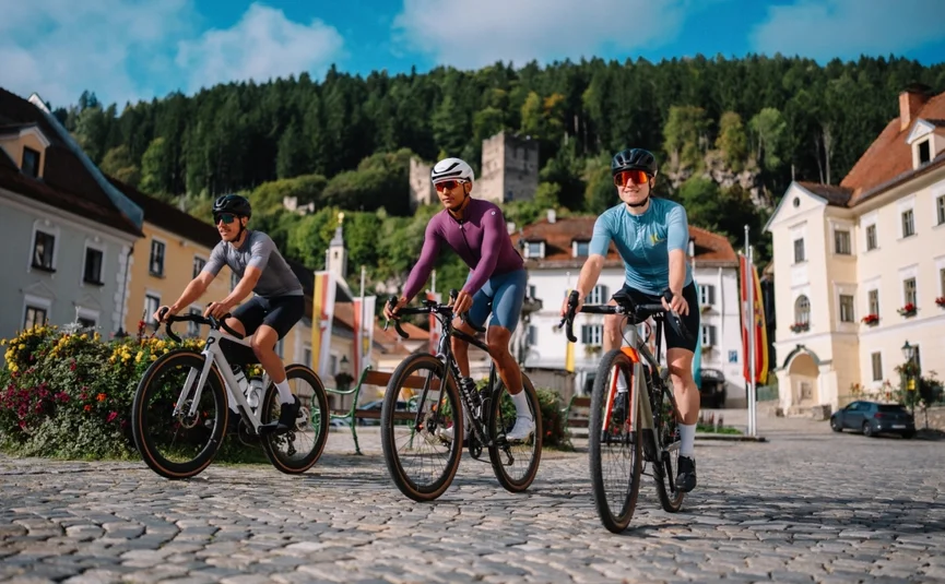 Drei Radfahrer auf Kopfsteinpflaster in einer Stadt mit Bergen im Hintergrund