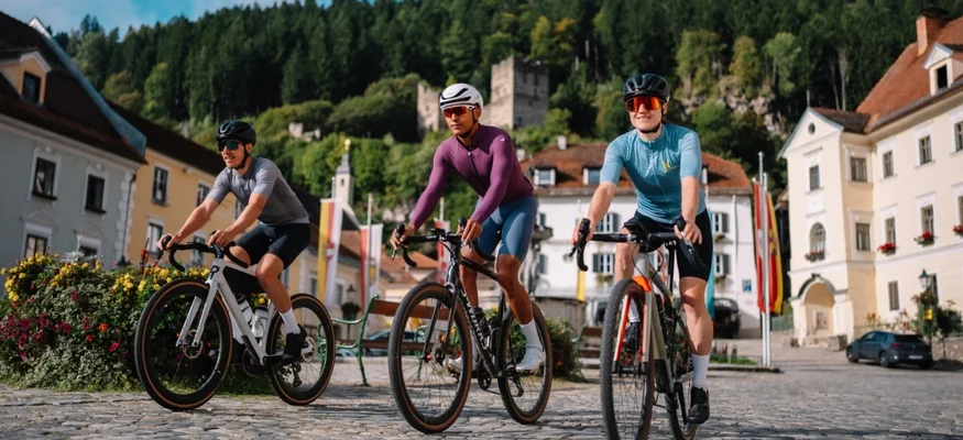 Drei Radfahrer auf Kopfsteinpflaster in einer Stadt mit Bergen im Hintergrund