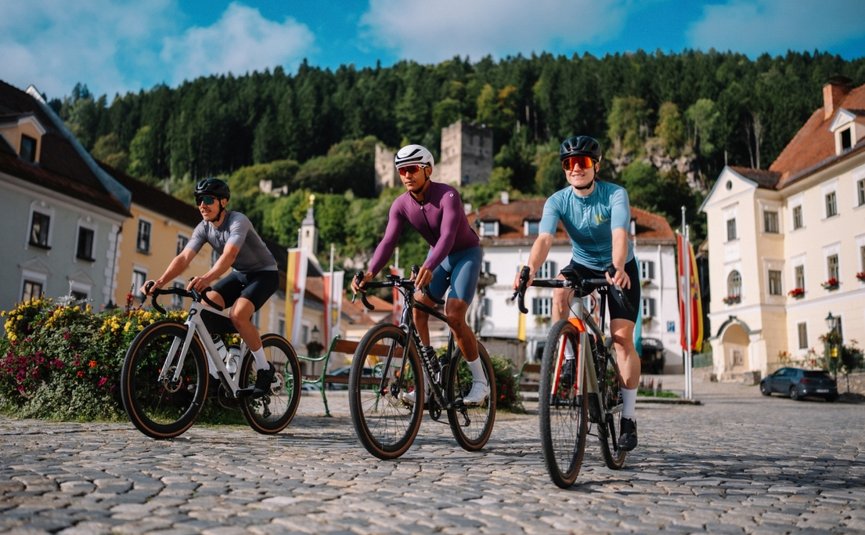 Stift St. Georgen **** © Martin Hoffmann Drei Radfahrer auf Kopfsteinpflaster in einer Stadt mit Bergen im Hintergrund