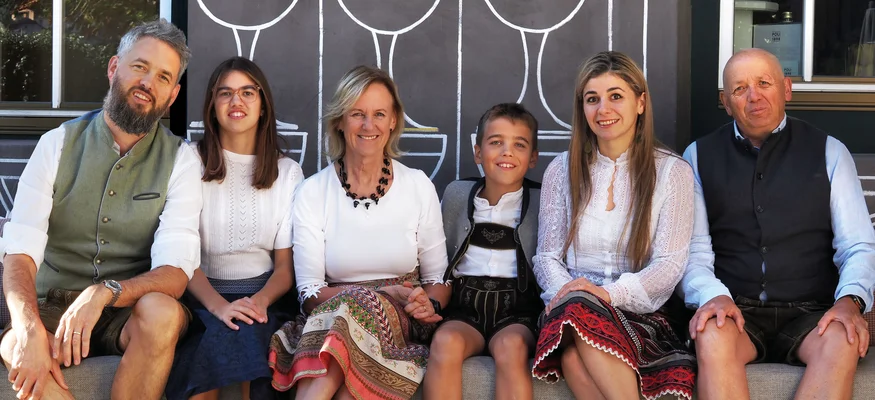 Gravel & Genuss am Kronplatz © Ueberall GmbH Familie in traditioneller Tracht sitzt auf einer Bank vor einer bemalten Wand