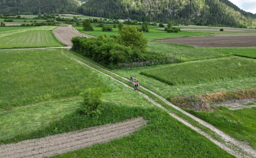 Zwei Radfahrer fahren auf einem Feldweg durch grüne Felder und Berge im Hintergrund