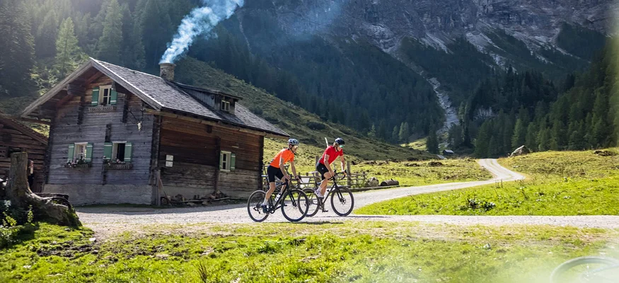 Salzburger Sportwelt © Salzburger Land Tourismus Zwei Radfahrer vor einer Hütte mit rauchendem Schornstein in den Bergen
