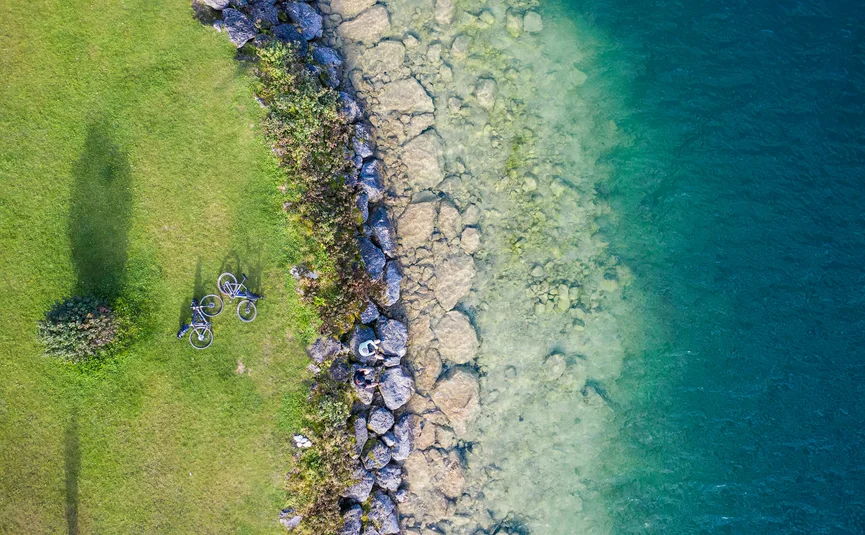 Two bicycles and people on rocks by clear lake next to green grass