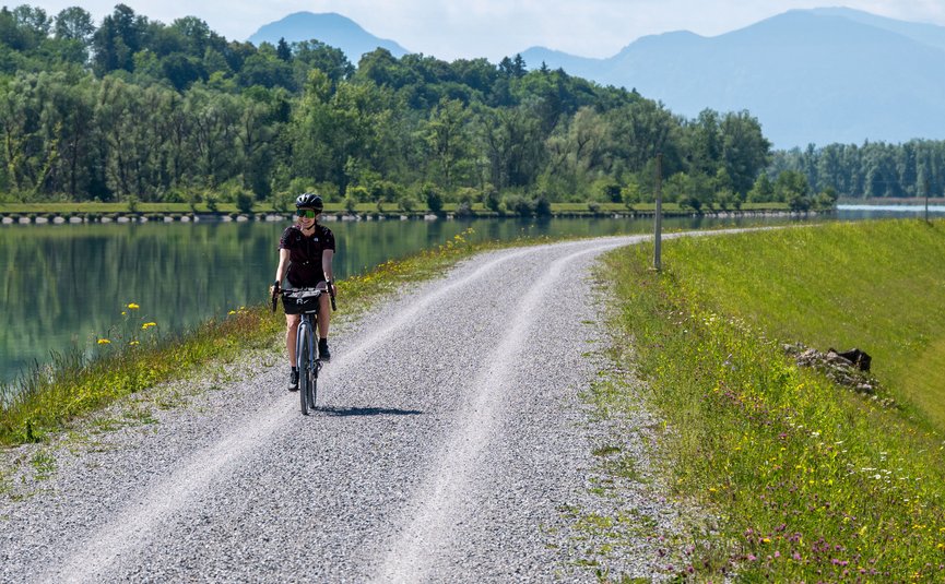 Fahrradfahrerin auf einem Schotterweg neben einem See und grünen Bäumen