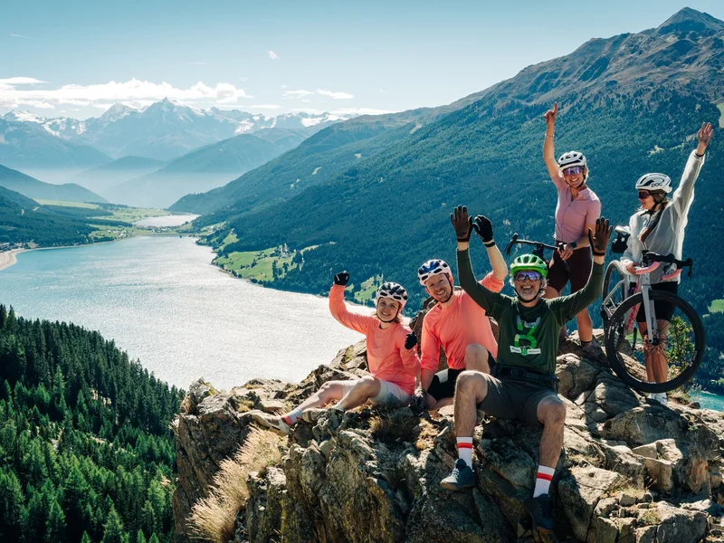 Gravelbike Holidays © TVB-Tiroler-Oberland-Nauders-Christian-Penning Eine Gruppe Radfahrer jubelt auf einem Felsen über einem türkisfarbenen Bergsee