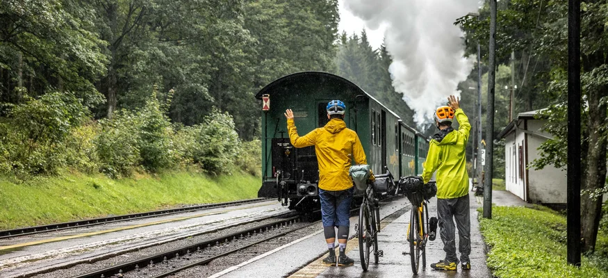 Zwei Radfahrer winken einem abfahrenden Dampflokzug im Wald zu