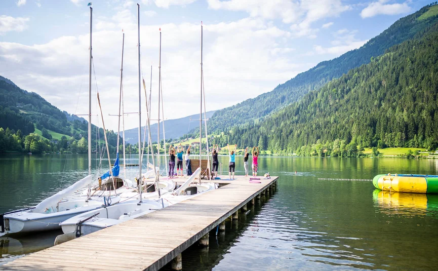 Menschen machen Yoga auf einem Steg am See mit Booten und Bergblick