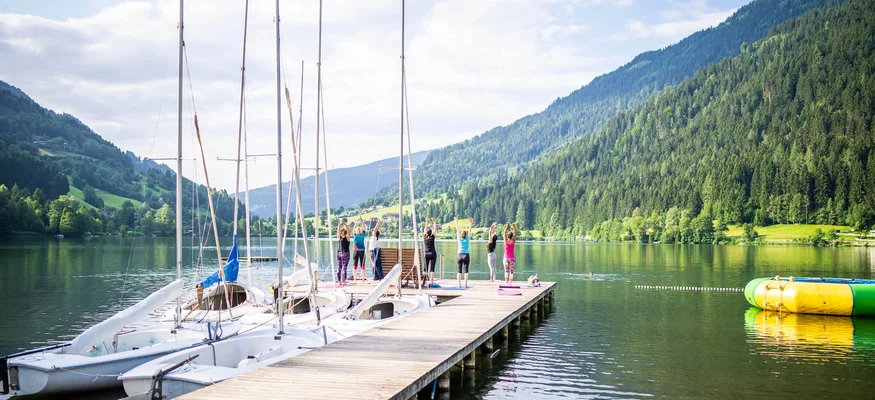 Menschen machen Yoga auf einem Steg am See mit Booten und Bergblick
