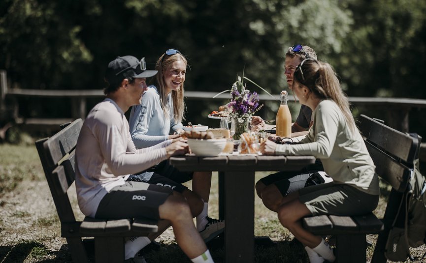 Four people sitting outdoors at a wooden table eating in the sunshine