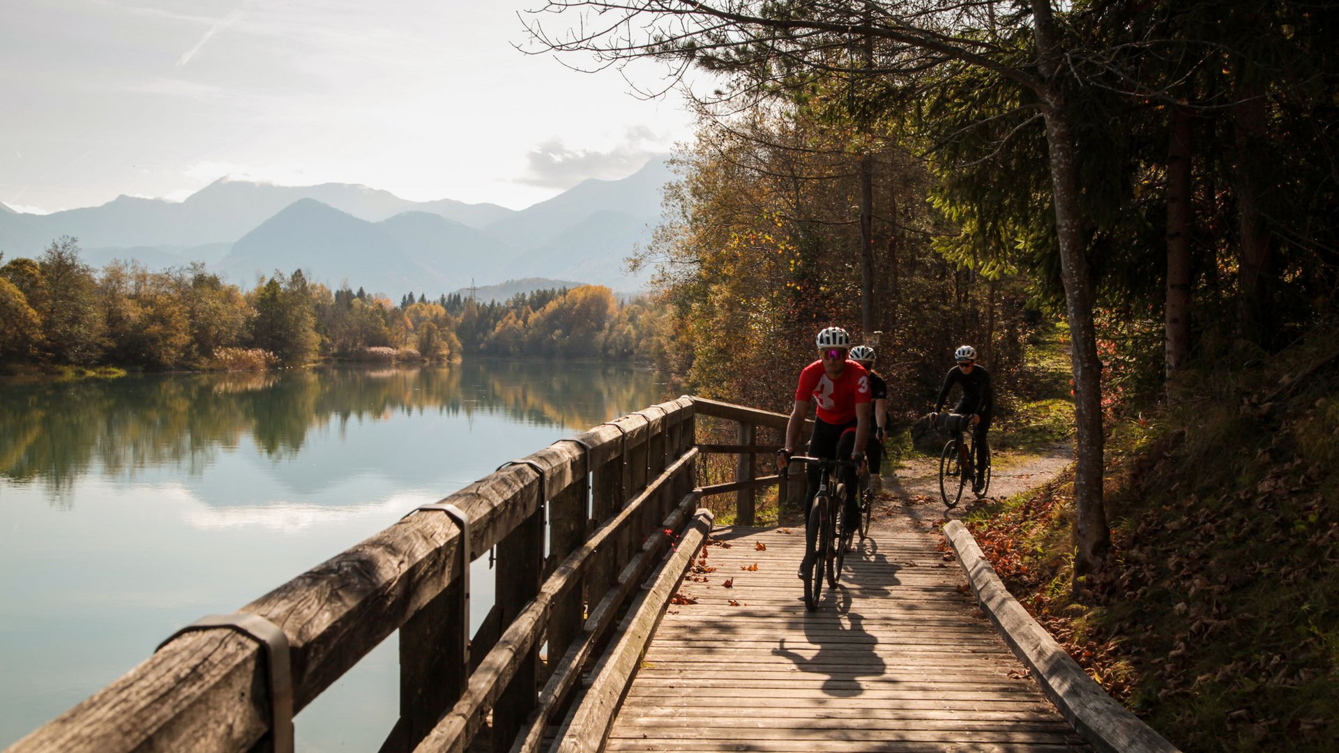 Drei Radfahrer fahren auf Holzsteg neben See in herbstlicher Landschaft
