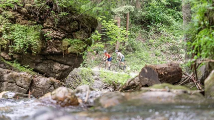 Zwei Radfahrer fahren auf Waldweg neben Bach im grünen Wald