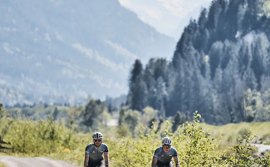 Zwei Radfahrer fahren auf einem Schotterweg in einer bergigen Landschaft.