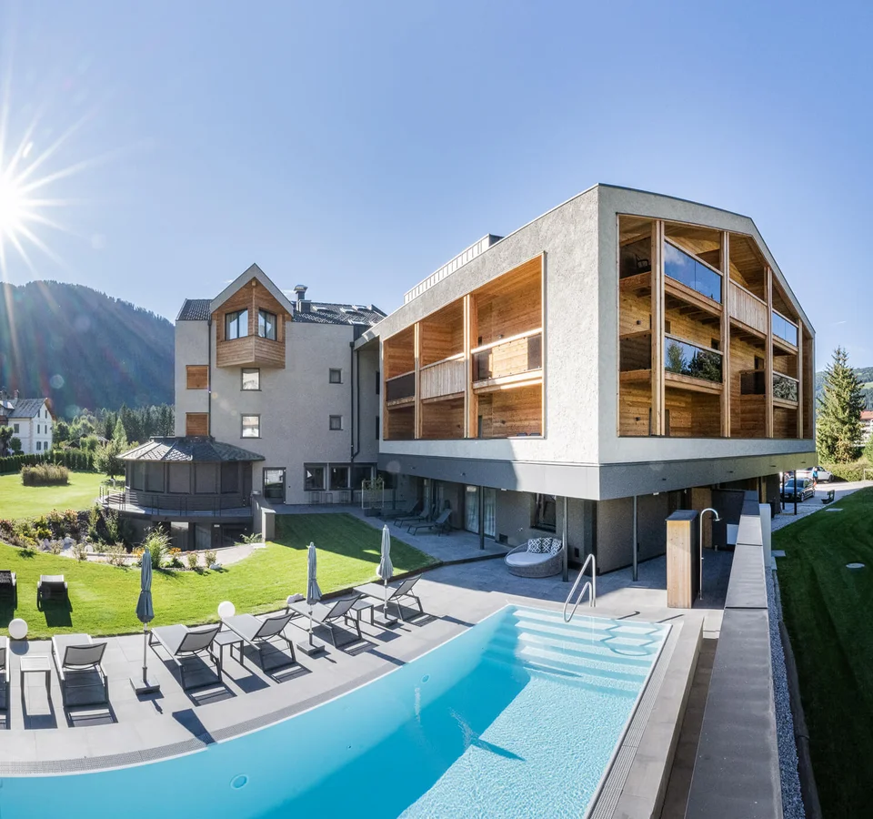 Modernes Hotel mit Pool und Sonnenliegen vor Bergkulisse bei sonnigem Himmel
