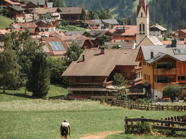 5 Gravelbike-Touren in Alta Badia, die du gefahren haben solltest © John Braynard Radfahrer fährt auf einem Weg durch eine Alpenlandschaft mit Dorf