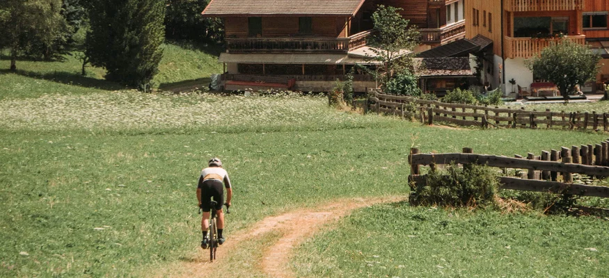 Gravelbiken ist eine Welt zwischendrin. © John Braynard Radfahrer fährt auf einem Weg durch eine Alpenlandschaft mit Dorf