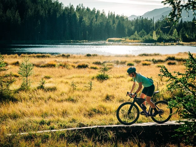 Das waren die Golden Days in Nauders © TVB-Tiroler-Oberland-Nauders-Christian-Penning - Gravelbiken in Nauders im Herbst Mountainbiker auf einem Holzsteg am See, umgeben von Wiesen, Wald und Bergen