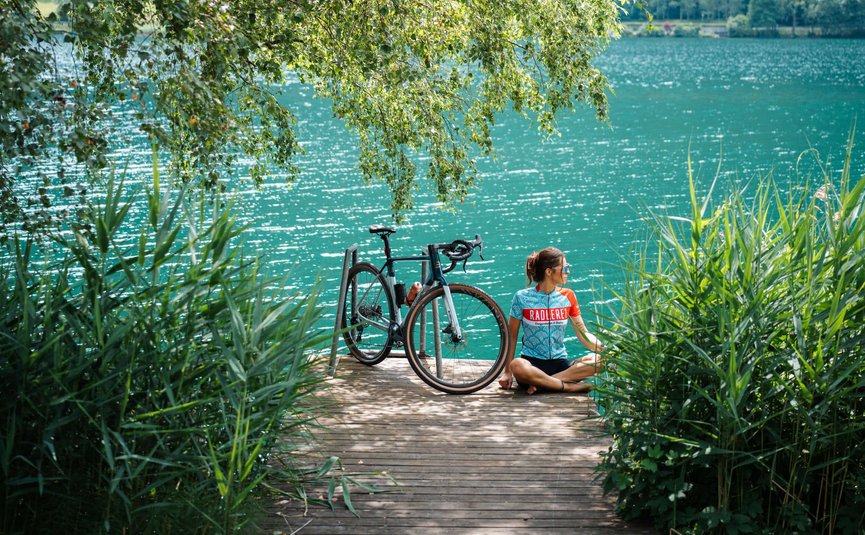 Woman sitting on dock next to bicycle by a lake with surrounding trees