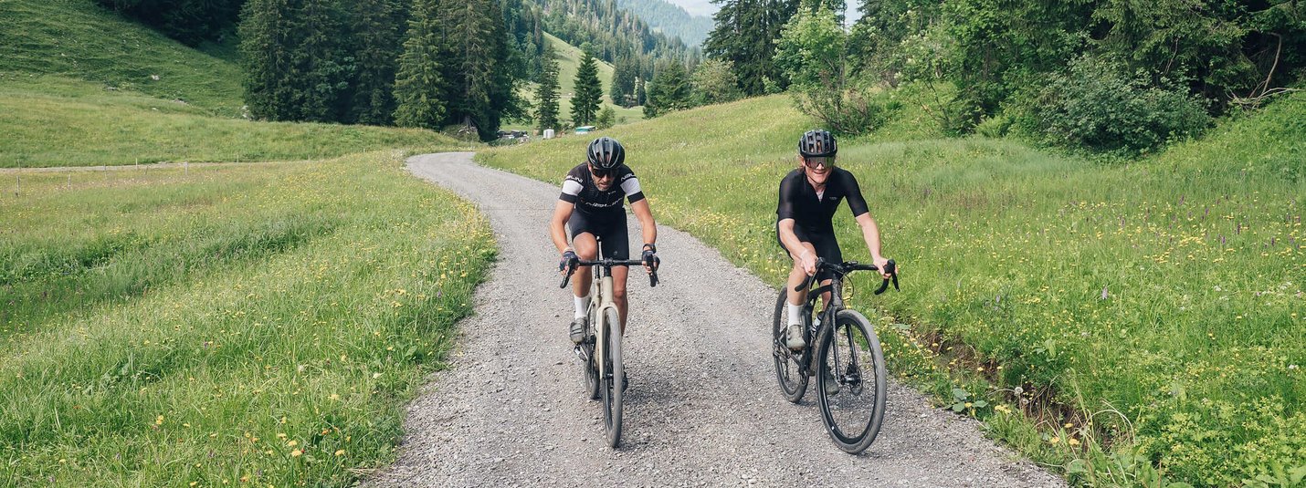 Two cyclists riding on a gravel path through a green mountain landscape