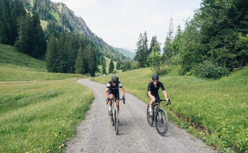 Two cyclists riding on a gravel path through a green mountain landscape