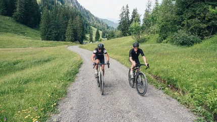 Zwei Radfahrer fahren auf einem Schotterweg durch eine grüne Berglandschaft