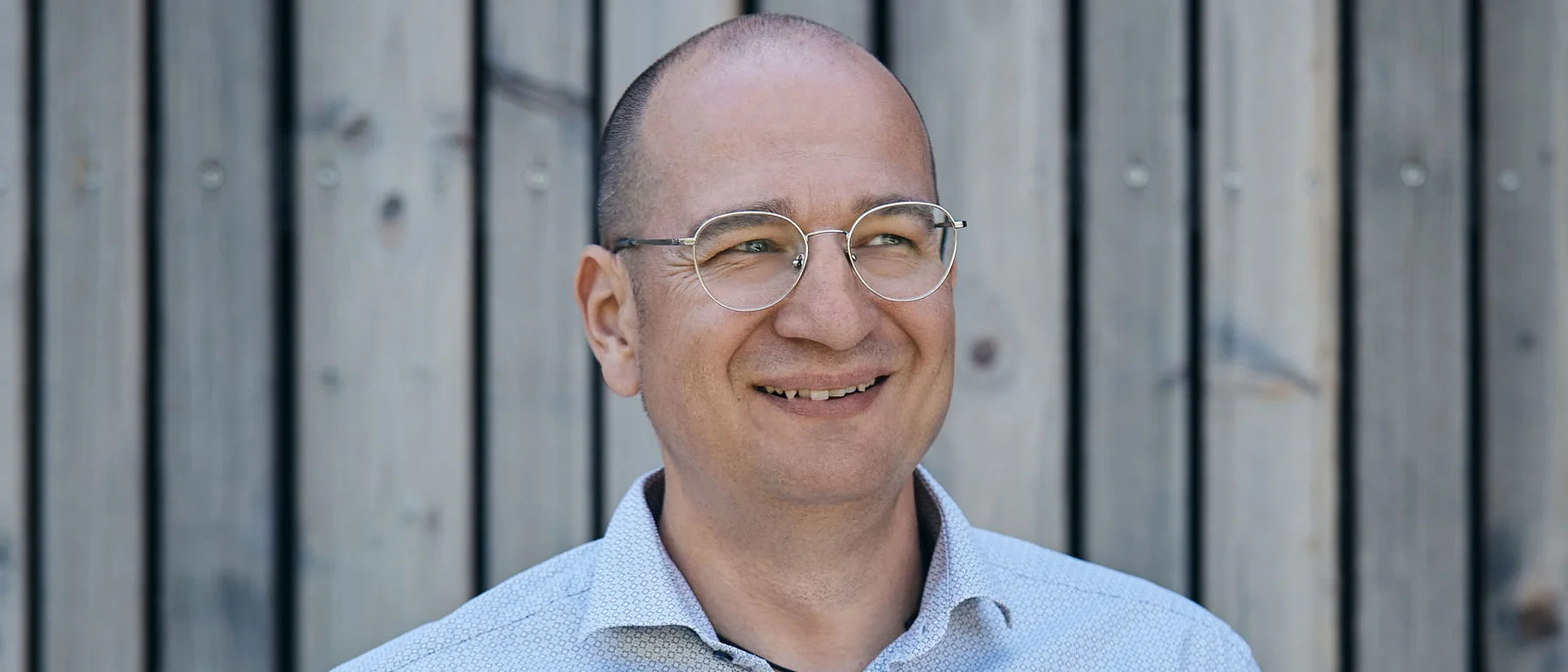 Lächelnder Mann mit Brille vor Holzwand trägt helles Hemd und Jeans