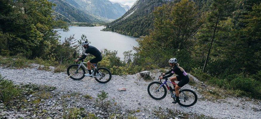 Zwei Radfahrer auf Bergweg mit See und Bergen im Hintergrund