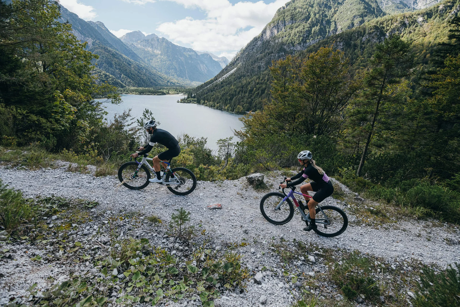 Gravelbike Holidays © WRTG Fotograf Sam Strauss Zwei Radfahrer auf Bergweg mit See und Bergen im Hintergrund