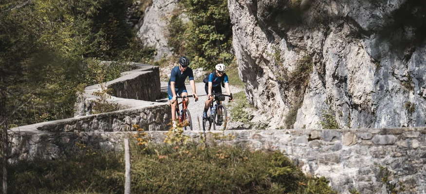 Zwei Radfahrer fahren auf einem Bergweg an Felsen und Bäumen vorbei