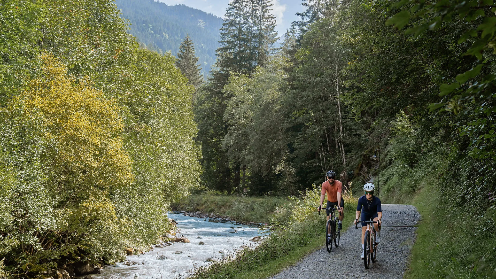 Grenzenlos graveln in Vorarlberg © Janine Brugger - Vorarlberg Tourismus Zwei Radfahrer auf einem Waldweg neben einem Fluss in den Bergen
