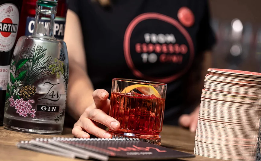 Hand holding a Negroni cocktail glass at a bar with gin and Campari bottles